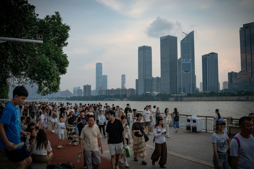 A crowd of people walking along a riverfront promenade with skyscrapers in the distance