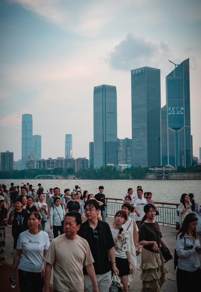 A crowd of people walking along a riverfront promenade with skyscrapers in the distance